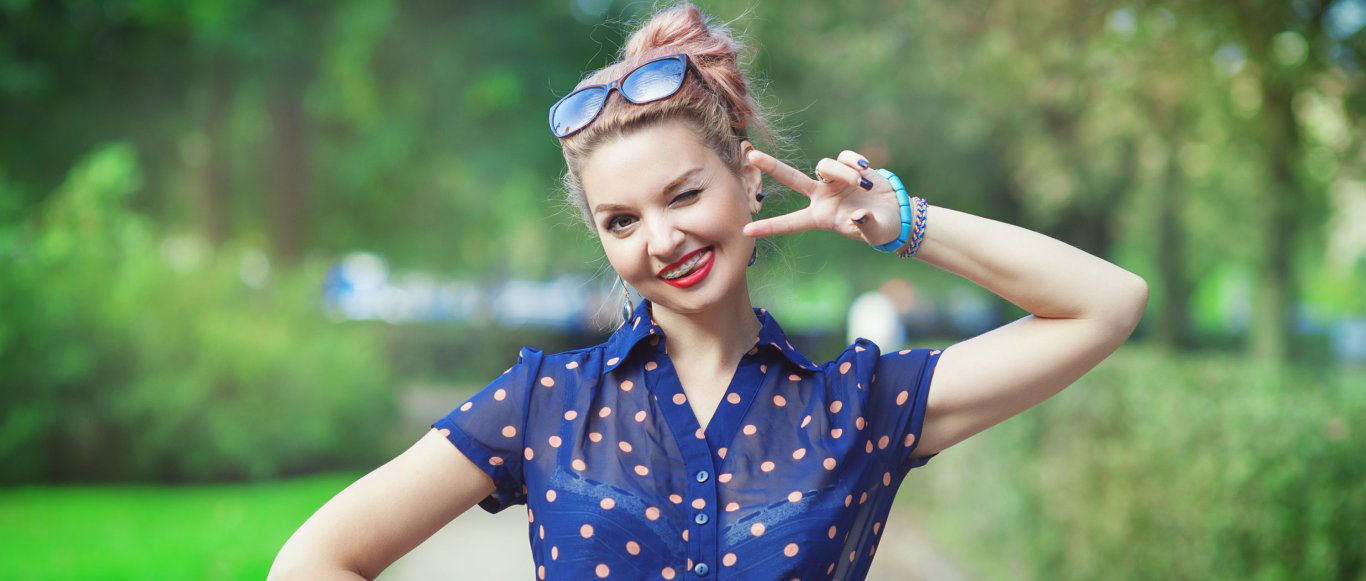 A woman poses with a peace sign gesture, wearing sunglasses, a polka dot shirt, and blue jeans.