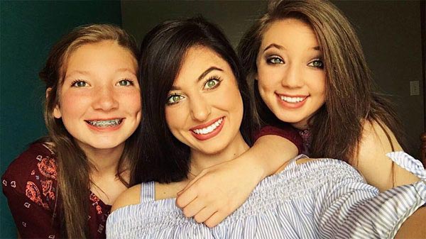 The image shows three individuals posing for a photograph with smiles on their faces  two girls are hugging each other while a woman stands behind them, all appear to be indoors with a blue wall visible in the background.