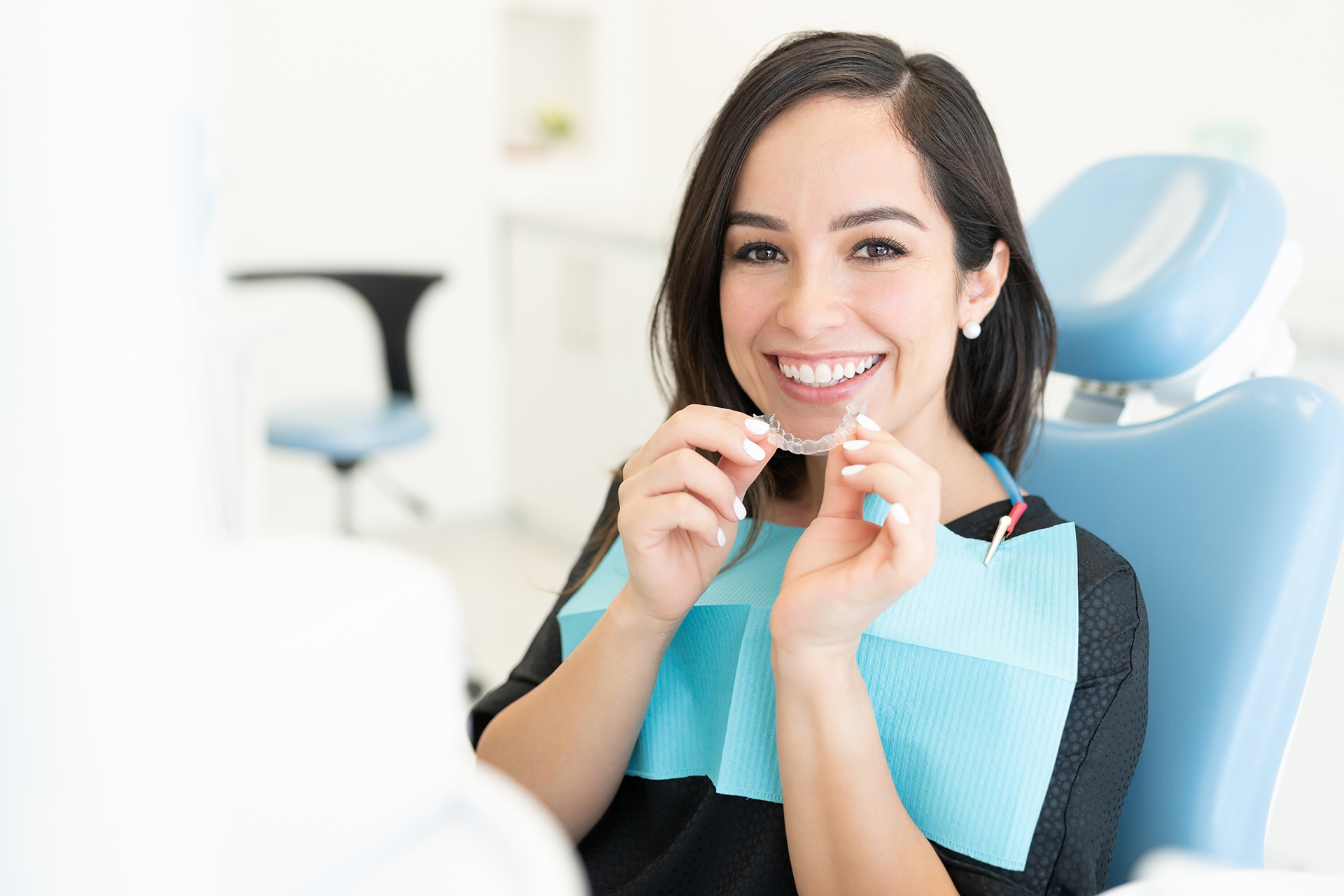 The image shows a woman sitting at a dental chair with a smile, holding up a toothbrush, wearing a blue apron, and surrounded by dental equipment.