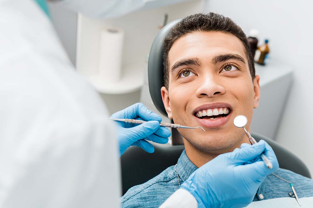 The image shows a person sitting in a dental chair with their mouth open wide, receiving dental treatment from a professional wearing a white coat and surgical mask, who appears to be in the process of cleaning or examining the person s teeth.