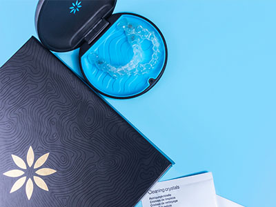 The image shows a cosmetic product with a blue lid and a patterned box placed on a surface next to a dark object with a white symbol, possibly a packaging item for skincare, set against a light background.
