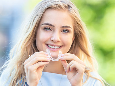 The image depicts a young woman smiling at the camera while holding a toothbrush with a toothpaste-covered bristle in her mouth, wearing a necklace, and standing outdoors during daylight.